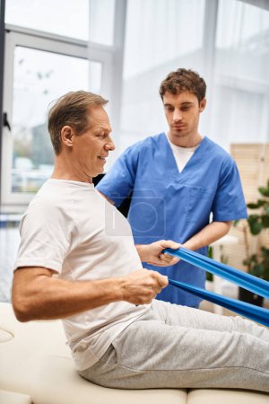 handsome dedicated doctor helping his mature jolly patient to use resistance band, rehabilitation