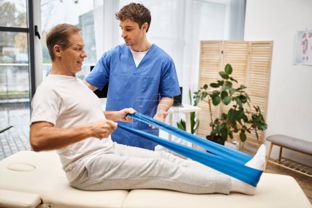 appealing devoted doctor helping his mature joyous patient to use resistance band, rehabilitation