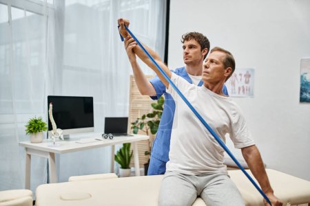 handsome dedicated doctor helping mature patient to use resistance band on rehabilitation in ward