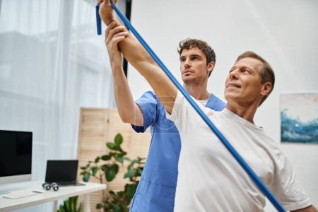 handsome doctor helping mature merry attractive patient to use resistance band on rehabilitation