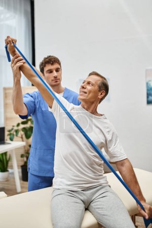 attractive doctor in blue robe helping mature jolly patient to use resistance band on rehabilitation