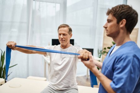 handsome devoted doctor in blue robe helping his mature jolly patient to use resistance band
