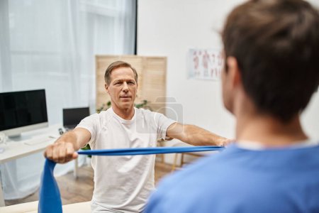 focus on joyous mature man in cozy attire using resistance band in front of his blurred doctor