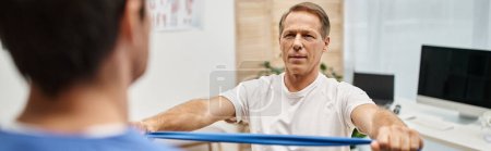 focus on jolly handsome mature man using resistance band in front of his blurred doctor, banner