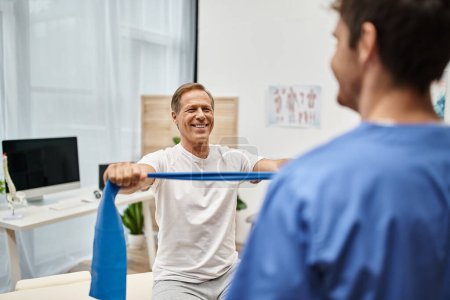 focus on cheerful mature man in casual attire using resistance band in front of his blurred doctor