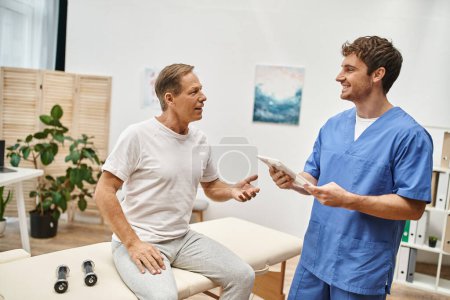 happy hardworking doctor with tablet talking to his mature cheerful patient during rehabilitation