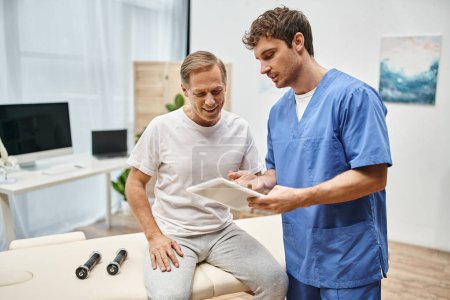 atractivo médico en bata azul sosteniendo tableta y hablando con su paciente maduro durante la cita