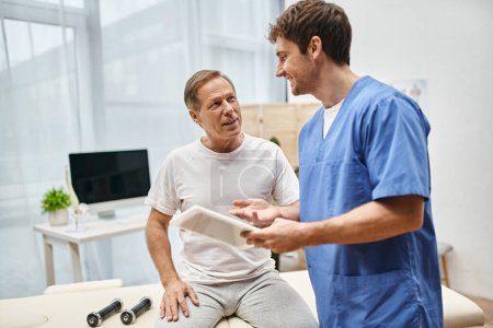 happy devoted doctor holding tablet and talking to his mature jolly patient during rehabilitation