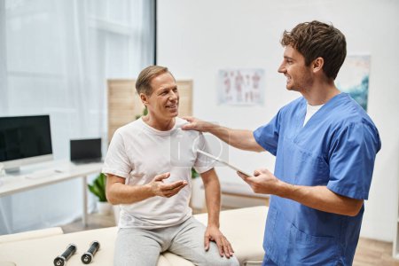 médico dedicado alegre sosteniendo la tableta y hablando con su paciente feliz madura durante la rehabilitación