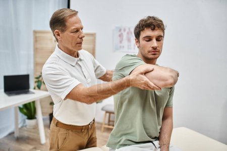 Photo for Handsome hardworking rehabilitologist helping his patient in casual attire to rehabilitate in ward - Royalty Free Image