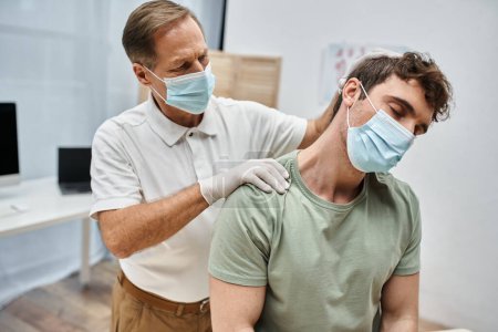 Téléchargez les photos : Docteur mature dévoué en uniforme avec masque massant le cou de son patient dans la salle d'hôpital - en image libre de droit
