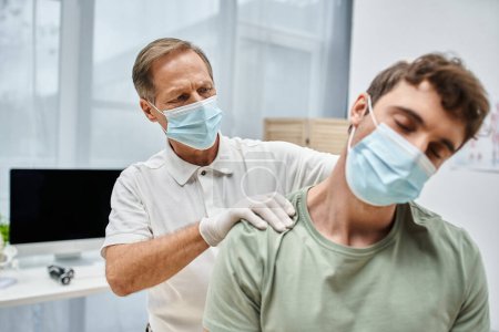Téléchargez les photos : Dévoué réhabilitologue mature avec masque et gants massant le cou de son patient dans la salle d'hôpital - en image libre de droit