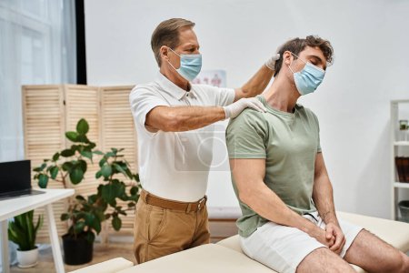 Photo for Devoted handsome mature doctor in uniform with mask massaging neck of his patient in hospital ward - Royalty Free Image