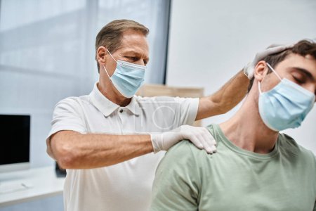 Photo for Hardworking mature doctor with mask and gloves massaging neck of his patient in hospital ward - Royalty Free Image