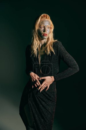 Téléchargez les photos : Jeune femme tatouée avec des paillettes sur le visage et les lèvres rouges posant en robe sur fond noir - en image libre de droit