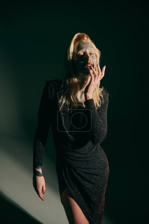 Téléchargez les photos : Femme blonde tatouée avec des paillettes sur le visage et les lèvres rouges posant en robe brillante sur fond noir - en image libre de droit