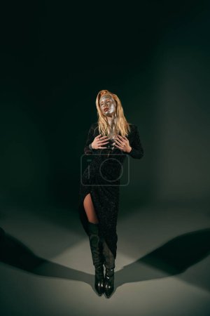 alluring woman with glitter on face posing in shiny dress and knee high boots on black background
