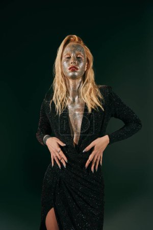 Téléchargez les photos : Femme blonde tatouée avec des paillettes sur le visage et le corps posant en robe brillante sur fond noir - en image libre de droit