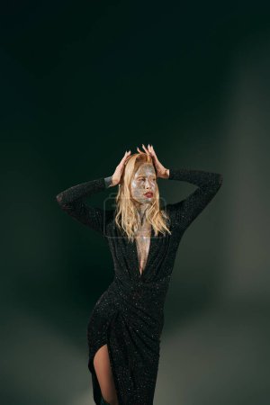 blonde woman with glitter on face posing in shiny dress with hands near hair on black backdrop