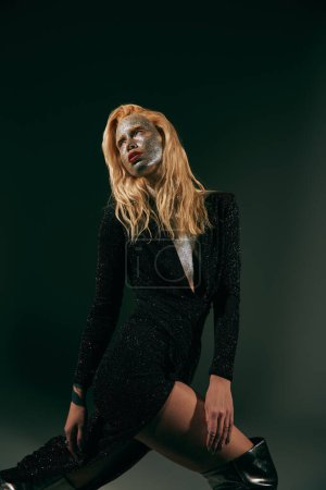young blonde woman with sparkling glitter on face posing in trendy shiny dress on black backdrop