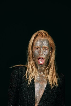 provokante junge blonde Frau mit Glitzerschminke, Zunge mit Pille auf schwarzem Hintergrund