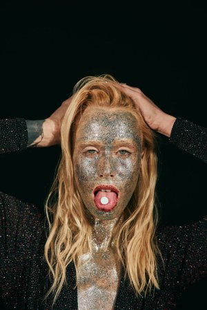 provokante junge blonde Frau mit Glitzern im Gesicht, Zunge mit Pille auf schwarzem Hintergrund