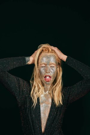 young blonde woman with glitter makeup and closed eyes showing tongue with pill on black background