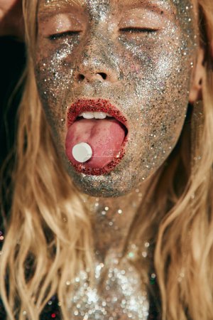 close up of young woman with glitter makeup and closed eyes showing tongue with pill on black