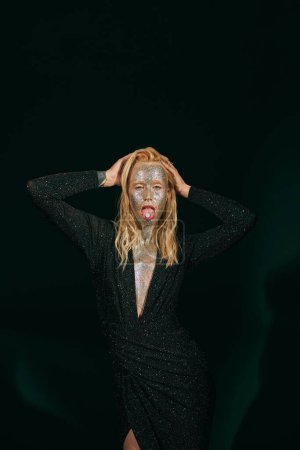 striking blonde woman with glitter makeup and closed eyes showing tongue with pill on black