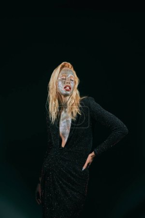 striking blonde woman with glitter makeup holding pill in teeth and posing with hand on hip on black