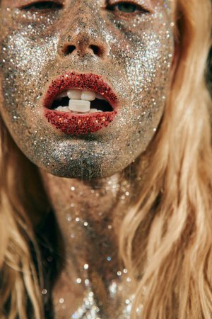 close up of blonde woman with glitter makeup holding pill in teeth and posing on black background