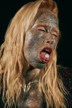 daring young woman with glitter makeup holding pill on tongue and posing with closed eyes on black