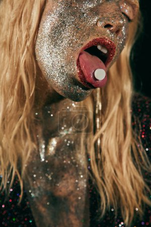 beschnitten wagemutige Frau mit Glitzer-Make-up hält Pille auf der Zunge und posiert auf schwarzem Hintergrund