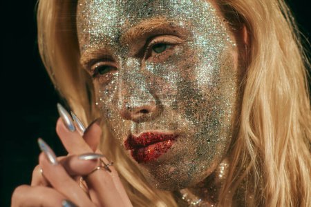 close up of blonde woman with green eyes and glitter on face and body posing on black backdrop