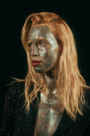 Porträt einer blonden Frau mit grünen Augen und Glitzern im Gesicht und Körper, die vor schwarzem Hintergrund posiert