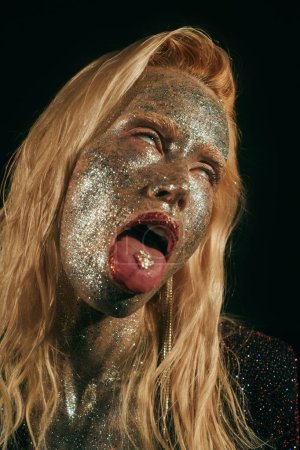 portrait of provocative blonde woman with glitter on face and body rolling eyes on black backdrop