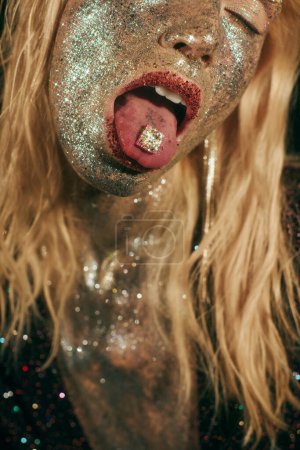 cropped shot of blonde woman with glitter on face and sparkling pill on tongue on black backdrop