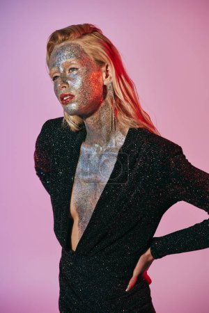 Téléchargez les photos : Femme blonde frappante avec maquillage paillettes et robe brillante posant avec les mains sur les hanches sur fond rose - en image libre de droit