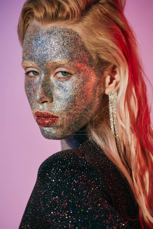 portrait of striking blonde woman with glitter makeup and shiny dress posing on pink backdrop