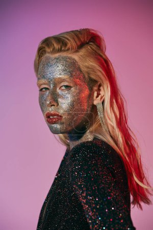 portrait of striking and blonde woman with glitter makeup and shiny dress posing on pink backdrop