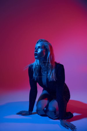 blonde and stylish  woman covered in glitter posing in shiny outfit under colored lights in studio