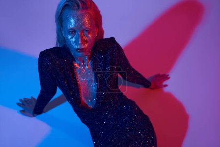 alluring sparkling model covered in glitter posing in shiny outfit under colored lights in studio