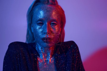 portrait of sparkling model covered in glitter posing in shiny outfit under colored lights in studio