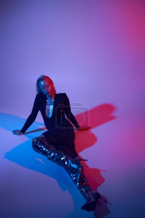 selbstbewusste junge Frau im Glitzerlook posiert in glänzendem Outfit unter neonfarbenen Lichtern im Studio