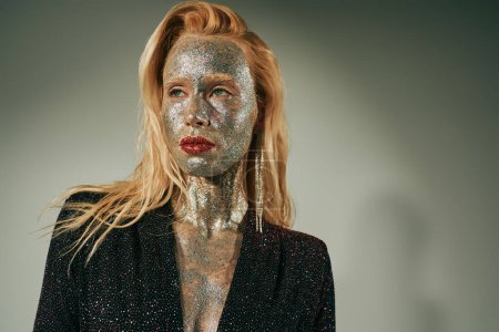 dreamy blonde woman in shiny dress with glitter all over her body and face posing on grey backdrop