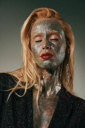 alluring young woman with closed eyes and glitter all over body and face posing on grey backdrop