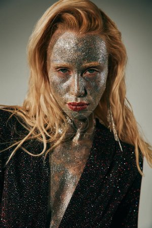Porträt einer blonden Frau mit grünen Augen und Glitzern am ganzen Körper und Gesicht, die vor grauem Hintergrund posiert