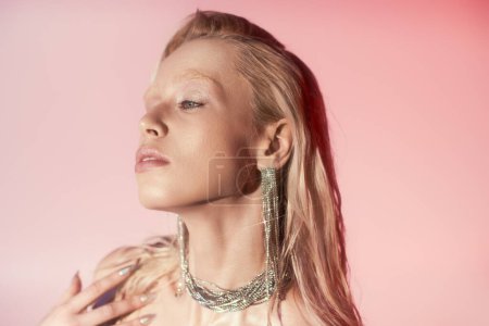serene beauty, young blonde woman in shiny necklace and earrings posing on pink background