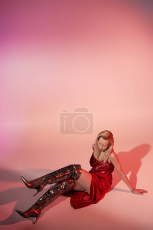 atractiva mujer de moda con el pelo largo y rubio en vestido rojo sentado en el suelo sobre fondo rosa