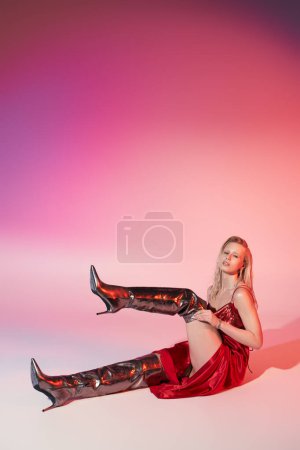 Photo for Alluring woman with blonde hair in red dress sitting on floor with raised leg and looking at camera - Royalty Free Image
